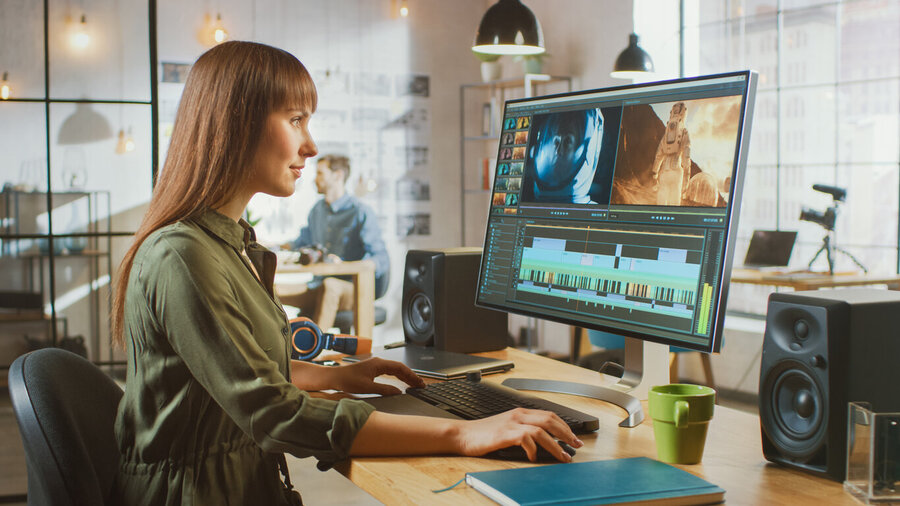 Beautiful Brunette Female Video Editor Works with Footage on Her Personal Computer with Big Display. She Works in a Cool Office Loft. Other Male Creative Colleague Walks in the Background.